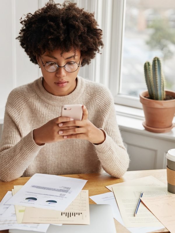 Busy businesswoman with Afro hairstyle, works with documentation, checks bank account via cell phone