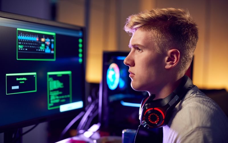 Male Teenage Hacker Sitting In Front Of Computer Screens Bypassing Cyber Security