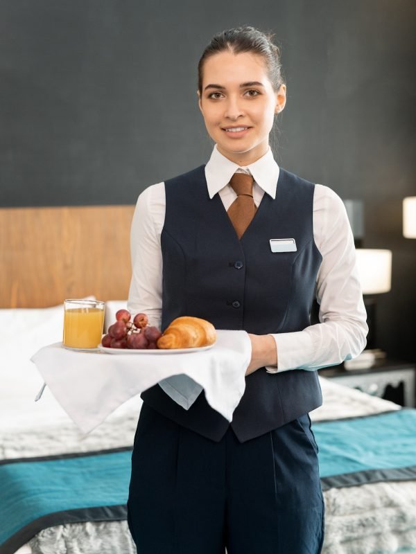 Happy young elegant female worker of hotel holding tray with breakfast against bed in hotel room