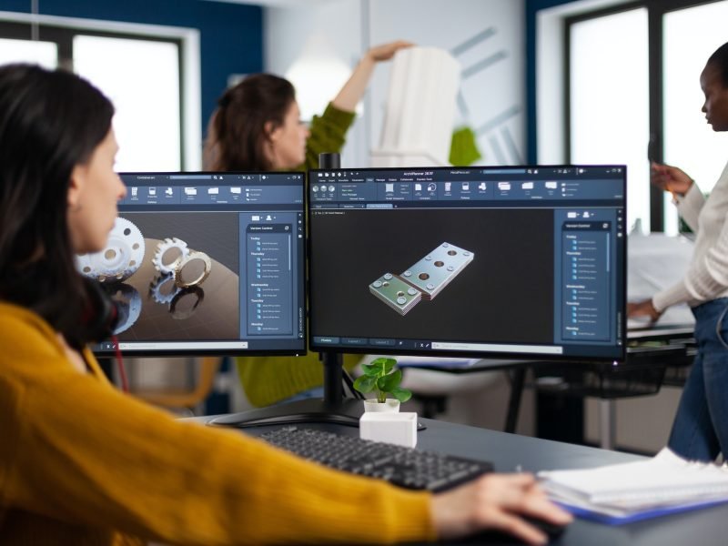 Industrial engineer woman working at pc with two monitors, screens showing CAD software