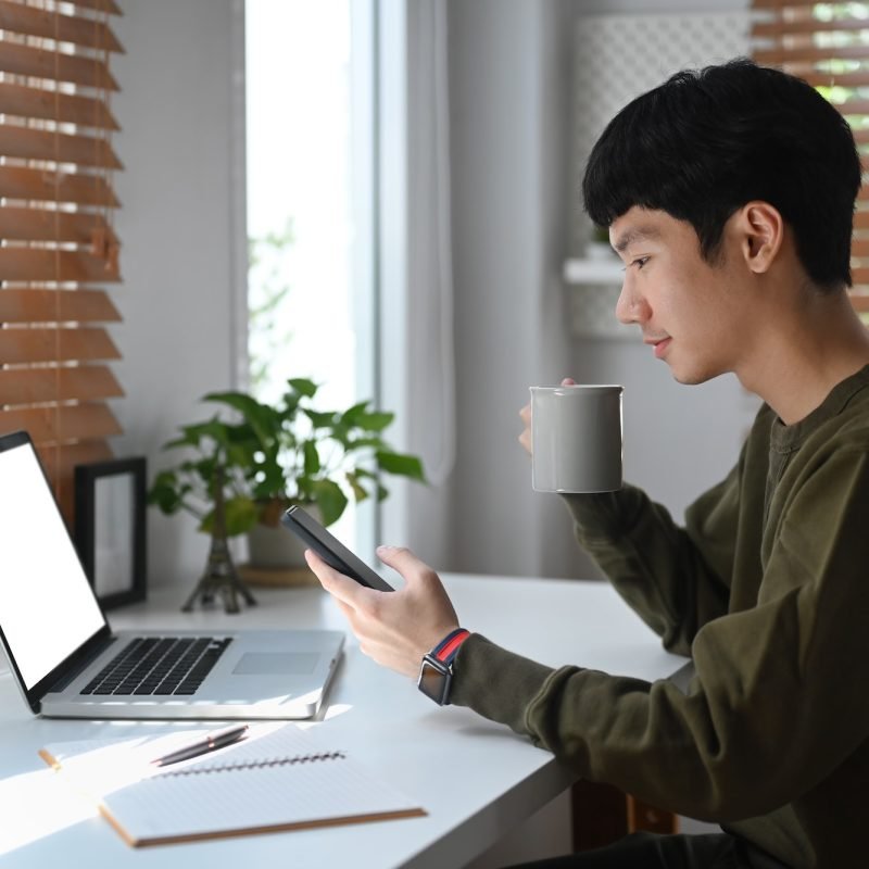 Side view of young businessman drinking coffee an checking social media on mobile phone.